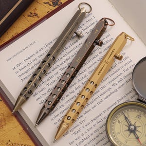 May include: Four metal pens in bronze and gold tones lie on a book. The pens feature a distinctive design with circular cutouts and a ring at the top. A compass and magnifying glass are also present.