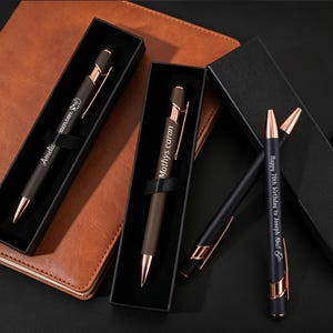 May include: A collection of matte black ballpoint pens with rose gold accents and clips. Two pens are housed in individual black gift boxes, while two others lie outside. The pens feature custom engravings including names and Happy 70th birthday to Joseph Abu!
