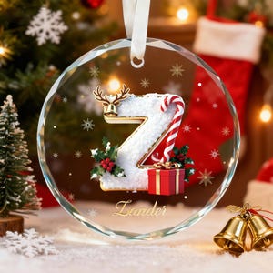 Puede incluir: Un adorno navideño de cristal transparente con un diseño decorativo de la letra "Z". La letra es blanca con un bastón de caramelo, acebo y una pequeña caja de regalo roja. El adorno tiene una campana de reno dorada y el nombre "Lander" en oro.