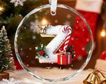 May include: A clear glass Christmas ornament with a decorative letter "Z" design. The letter is white with a candy cane, holly, and a small red gift box. The ornament has a gold reindeer bell and the name "Lander" in gold.