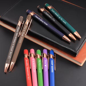 Op de afbeelding: Een assortiment gepersonaliseerde pennen in diverse kleuren, waaronder bruin, rood, roze, groen, paars en blauw, met goudkleurige accenten. De pennen zijn gegraveerd met teksten als "Happy 70th birthday to Joseph Abul", "Mia & Book 2025" en andere namen en data. De pennen liggen op een zwart en bruin notitieboek.