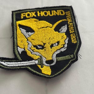 Może przedstawiać: Haftowana naszywka z ż&oacute;łtą głową lisa, białymi detalami i nożem. Naszywka zawiera napisy "FOX HOUND" i "SPECIAL FORCE GROUP" w kolorze zielonym i białym na czarnym tle. Idealna na kurtki lub torby.