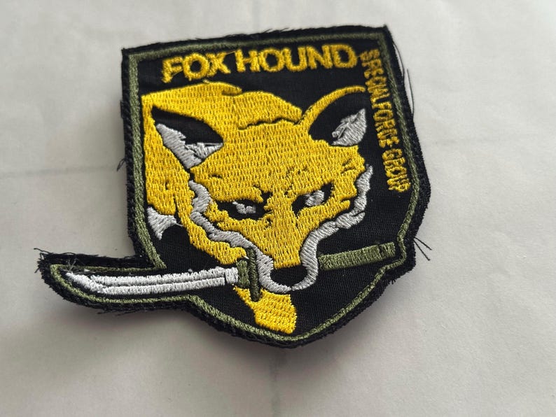 Może przedstawiać: Haftowana naszywka z ż&oacute;łtą głową lisa, białymi detalami i nożem. Naszywka zawiera napisy "FOX HOUND" i "SPECIAL FORCE GROUP" w kolorze zielonym i białym na czarnym tle. Idealna na kurtki lub torby.