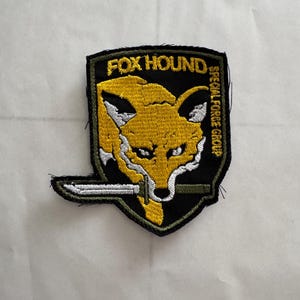 May include: Embroidered patch featuring a yellow fox head with white accents, holding a knife in its mouth. The patch has the text "FOX HOUND" and "SPECIAL FORCE GROUP" on a black background.
