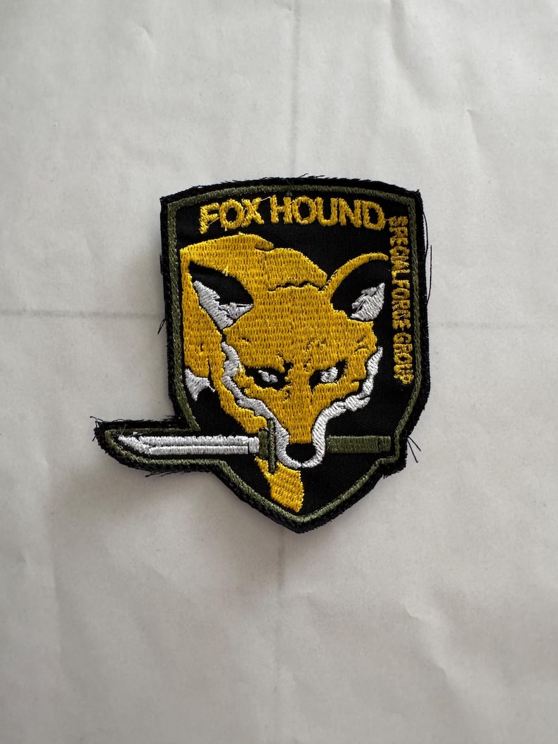 Może przedstawiać: Haftowana naszywka z ż&oacute;łtą głową lisa, białymi detalami i nożem w pysku. Na naszywce widnieje napis "FOX HOUND" i "SPECIAL FORCE GROUP" na czarnym tle. Emblemat w stylu militarnym.