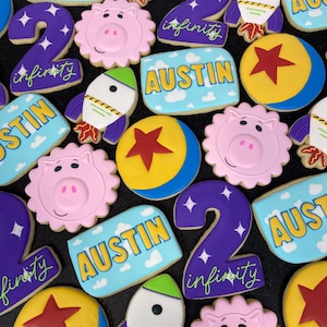 May include: A collection of decorated sugar cookies featuring a Toy Story theme. The cookies are decorated with characters like Buzz Lightyear, Woody, and the alien. The cookies also feature the name "Austin" and the number "2" with the word "infinity" written on them.