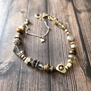May include: A bracelet featuring a variety of metallic, glass, and crystal beads in shades of gold, bronze, and silver. The design includes heart-shaped charms, faceted beads, and a delicate silver-toned chain extension with a small heart pendant.