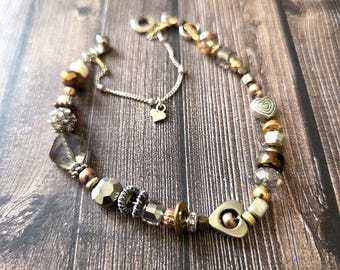 May include: A bracelet featuring a variety of metallic, glass, and crystal beads in shades of gold, bronze, and silver. The design includes heart-shaped charms, faceted beads, and a delicate silver-toned chain extension with a small heart pendant.
