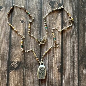 May include: A silver-toned necklace featuring a chain with interspersed small multi-colored beads in shades of teal, gold, brown, and metallic grey. The necklace includes a toggle clasp and a central silver-toned pendant with an embossed scrollwork design.