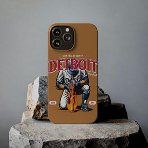 Könnte beinhalten: Eine braune iPhone-Hülle mit einem Baseballspieler-Motiv. Das Motiv zeigt einen Spieler in einer weißen und blauen Uniform, der einen Baseballschläger hält, der in Flammen steht. Der Text "CITY FULL OF GRITTY DETROIT Baseball ESTD 1894" ist auf der Hülle aufgedruckt.