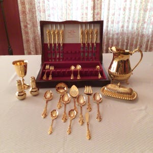 May include: A gold-plated flatware set in a wooden box with a red velvet lining. The set includes knives, forks, spoons, a serving spoon, a butter knife, a sugar spoon, a gravy ladle, a pitcher, a salt shaker, a pepper shaker, and a butter dish.
