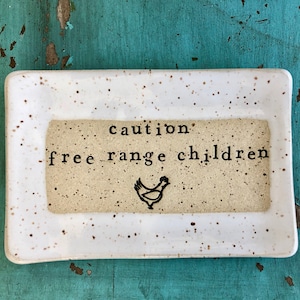 May include: White ceramic dish with brown speckles. The dish has a beige rectangle in the center with black text that reads "caution: free range children" and a black silhouette of a chicken.