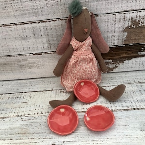 May include: A handmade plush bunny doll with long ears and a floral dress, accompanied by three small, round, coral-colored ceramic dishes. The bunny has a green pom-pom on its head. The dishes have a heart-shaped detail.