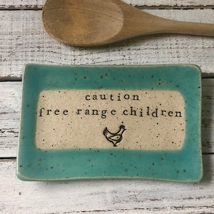 May include: A turquoise ceramic dish with a white interior and black text that reads "caution free range children" with a small black chicken silhouette.