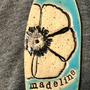 May include: A light blue ceramic tile with a white flower design and the name "madeline" written in black lettering.