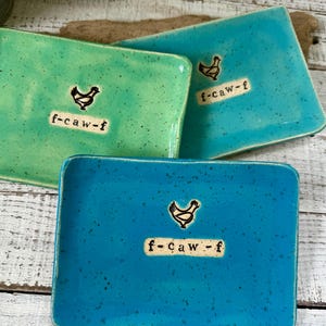 May include: Three square ceramic dishes in shades of teal and turquoise. Each dish features a black chicken illustration and the text "f-caw-f". The dishes are arranged on a rustic wooden surface, showcasing a farmhouse aesthetic.