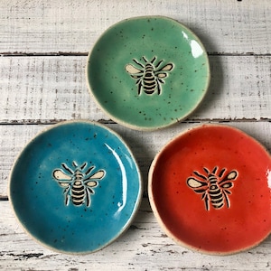 May include: Three small, round ceramic dishes in teal, red, and green, each featuring a bee design. The bee is carved into the center of each dish. The dishes have a speckled texture and a light-colored rim. The dishes are approximately 4 inches in diameter.