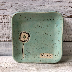 May include: A square ceramic dish with a speckled turquoise glaze. The dish has a black and white dandelion design and the word "wish" written on the bottom.