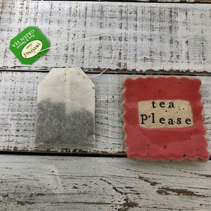 May include: A green tea bag with a green label that says "Twinings Green Tea" and a red ceramic dish with the text "tea please" on it.