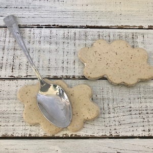 May include: Two beige ceramic spoon rests shaped like clouds. A silver spoon is resting on one of the spoon rests.