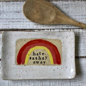 May include: A white ceramic dish with a speckled glaze features a rainbow design in red and yellow with the text "hate, sashay away" in black.