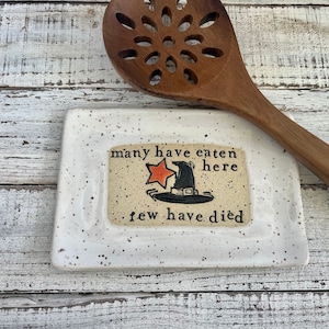 May include: White ceramic spoon rest with black and orange Halloween-themed design. The design features a witch's hat with a star and the text "many have eaten here few have died".