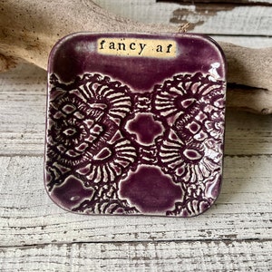 May include: A square, ceramic dish in a deep purple hue, featuring a raised, intricate white lace-like pattern. The words "fancy af" are printed in black on a beige rectangle. The dish is set against a rustic, white-washed wooden surface.