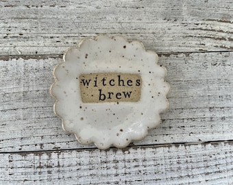 May include: A white ceramic dish with a scalloped edge and a speckled glaze. The dish has a small rectangular label with the text "witches brew" in black lettering.