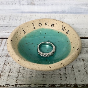 May include: A small, white ceramic ring dish with a turquoise blue glaze and speckles. The dish has the words "i love us" written on the rim. A silver ring with clear stones is sitting in the dish.