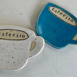 May include: Two ceramic coffee cup shaped dishes, one white with black speckles and the other blue with black speckles. Both dishes have the word "cafecito" written on them.