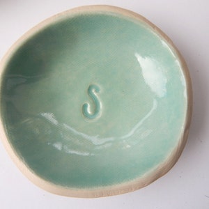 May include: A small, light blue ceramic dish with a raised letter "S" in the centre. The dish has a slightly irregular, handmade shape.