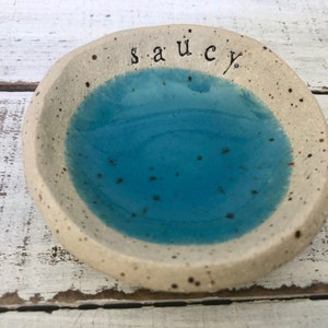 May include: A handmade ceramic dish with a speckled, off-white exterior and a vibrant turquoise interior. The word "saucy" is written in black script across the top. This small, round dish is perfect for holding small items.