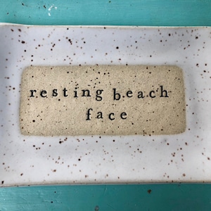 May include: White ceramic tray with brown speckles. The tray has a smaller rectangle in the center with the text "resting beach face" in black.