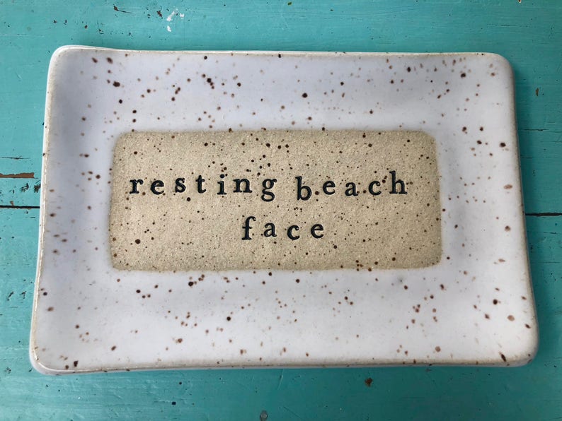 May include: White ceramic tray with brown speckles. The tray has a smaller rectangle in the center with the text "resting beach face" in black.