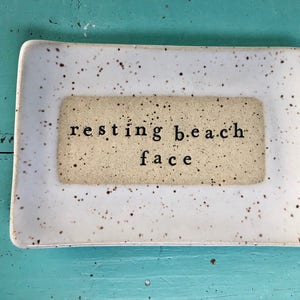May include: White ceramic dish with brown speckles and a beige rectangle in the center. The rectangle has the text "resting beach face" in black.