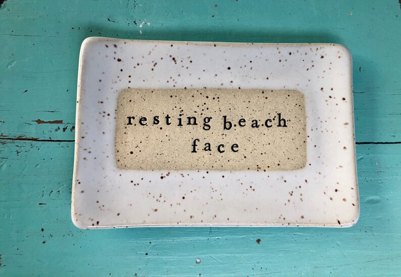 May include: White ceramic dish with brown speckles and a beige rectangle in the center. The rectangle has the text "resting beach face" in black.