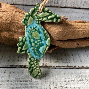 May include: A green and blue ceramic seahorse ornament with a crackled glaze finish. The ornament is hanging from a twine string.
