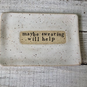 May include: A white ceramic dish with brown speckles. The dish has a rectangular shape and a small rectangular label with the text "maybe swearing will help" in black.