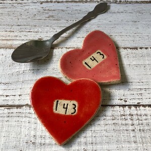 May include: Two red heart-shaped ceramic dishes with the number "143" written on them. The dishes are on a white wooden surface.