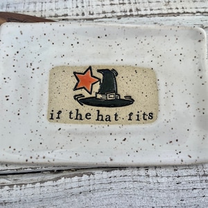 May include: A white ceramic dish with black speckles. The dish has a small rectangle in the center with a black witch hat with an orange star on top. The text "if the hat fits" is written below the hat.