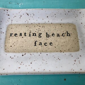 May include: White ceramic tray with brown speckles. The tray has a rectangular inset with the text "resting beach face" in black.