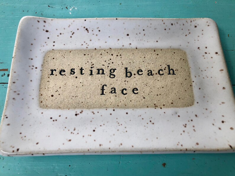 May include: White ceramic tray with brown speckles. The tray has a rectangular inset with the text "resting beach face" in black.