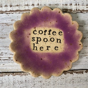 May include: A ceramic dish with a purple and white glaze. The dish has a scalloped edge and says "coffee spoon here" in black lettering.