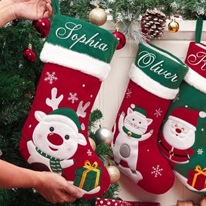 以下が含まれることがあります： 白いフェイクファーのトリムと緑色の吊り下げループが付いた赤いクリスマスストッキング。各ストッキングには、トナカイ、猫、サンタ、犬、ペンギンなど、さまざまなお祝いのキャラクターが描かれています。名前は緑色の袖口に刺繍されています。