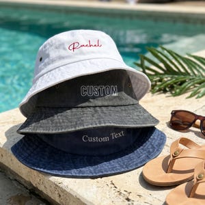May include: A stack of three bucket hats. The top hat is white with the red embroidered name Rachel. The middle hat is charcoal grey with the white embroidered word CUSTOM. The bottom hat is navy blue with the white embroidered text Custom Text.