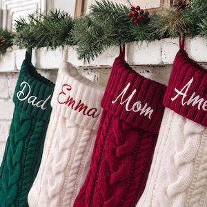 May include: Four knitted Christmas stockings featuring a cable knit pattern. The first is dark green with white embroidery reading Dad, the second is white with red embroidery reading Emma, the third is red with white embroidery reading Mom, and the fourth is white with red embroidery reading Amelia.