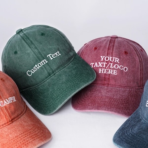 Könnte beinhalten: Vier Baseballkappen in den Farben Orange, Grün, Burgunderrot und Blau. Jede Kappe hat eine individuelle Textstickerei in Weiß. Die grüne Kappe trägt die Aufschrift "Custom Text", die bordeauxfarbene "YOUR TEXT/LOGO HERE", die orangefarbene "HAPPY CAMPER" und die blaue "Custom Embroidery".