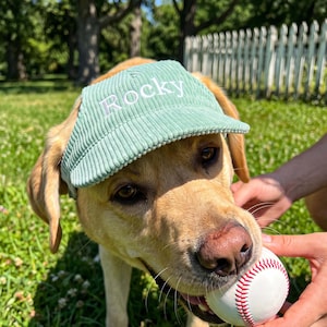 May include: A light green corduroy baseball cap featuring the name Rocky embroidered in white thread on the front panel. The cap has a structured brim and a textured fabric surface, designed to fit over the head of a dog.