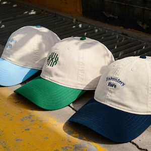 May include: Three baseball caps with different colored brims. The first cap has a light blue brim and the text "Downtown Girl" embroidered on the front. The second cap has a green brim and the text "XXX" embroidered on the front. The third cap has a dark blue brim and the text "Embroidery Here" embroidered on the front.
