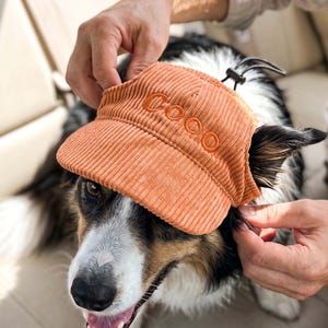 May include: An orange corduroy pet visor featuring the name Coco embroidered in matching orange thread on the front panel. The visor includes an adjustable black elastic cord with a plastic toggle fastener to secure it around the head.